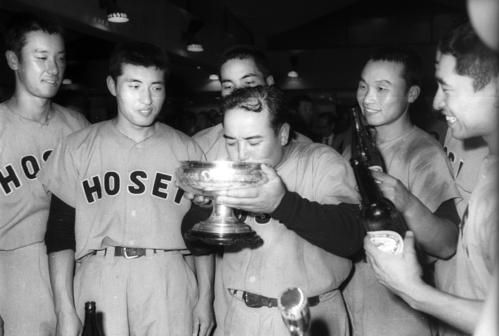 62年　優勝し、ナインが見つめる前で天皇杯に注がれた美酒を飲み干す法大・田丸仁監督（中央）