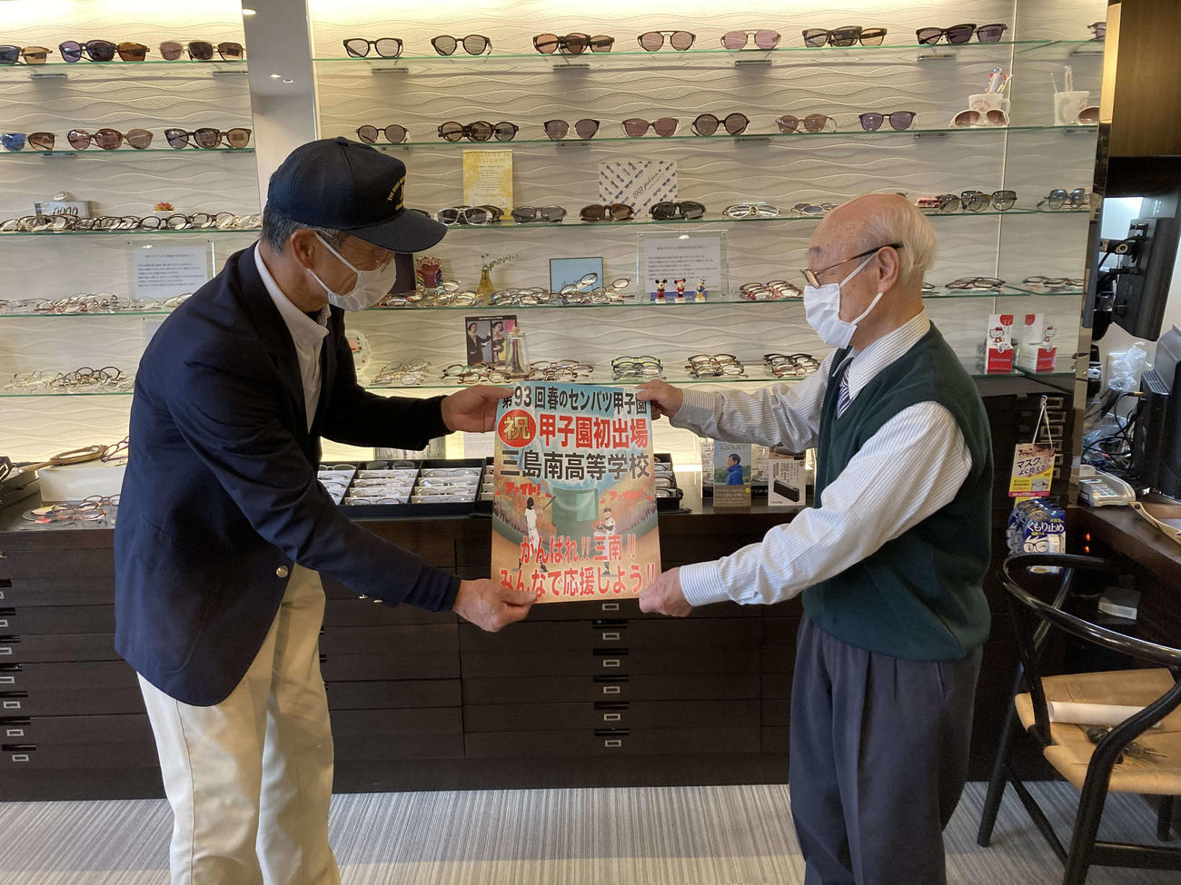 三島南OB会長の諏訪部孝志さん（左）は、ポスターを持って地元商店街を回った。眼鏡店ではポスター掲示と寄付に快く応じてくれた（諏訪部さん提供）