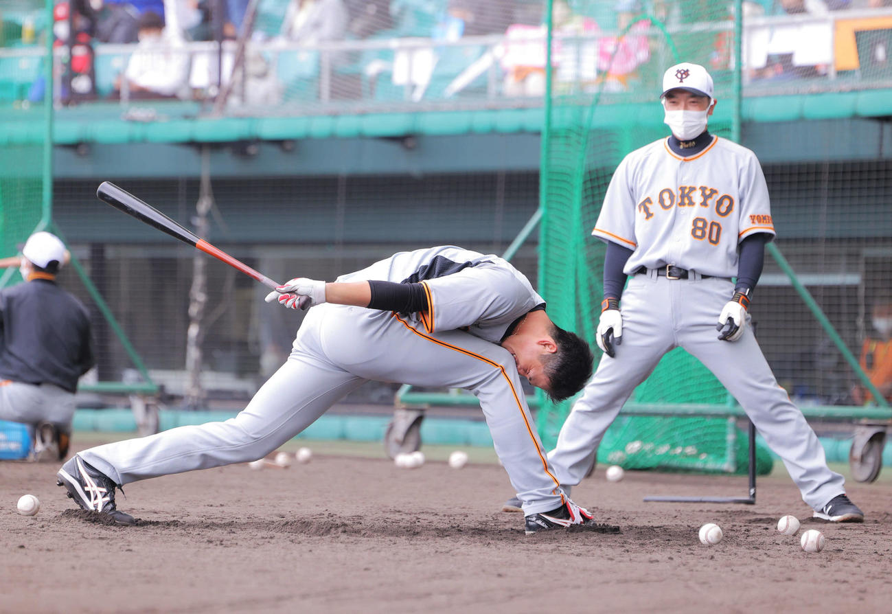 阿部作戦兼ディフェンスチーフコーチ（右）の前で、険しい表情で関節を伸ばしながらトス打撃に臨む巨人秋広（撮影・河田真司）