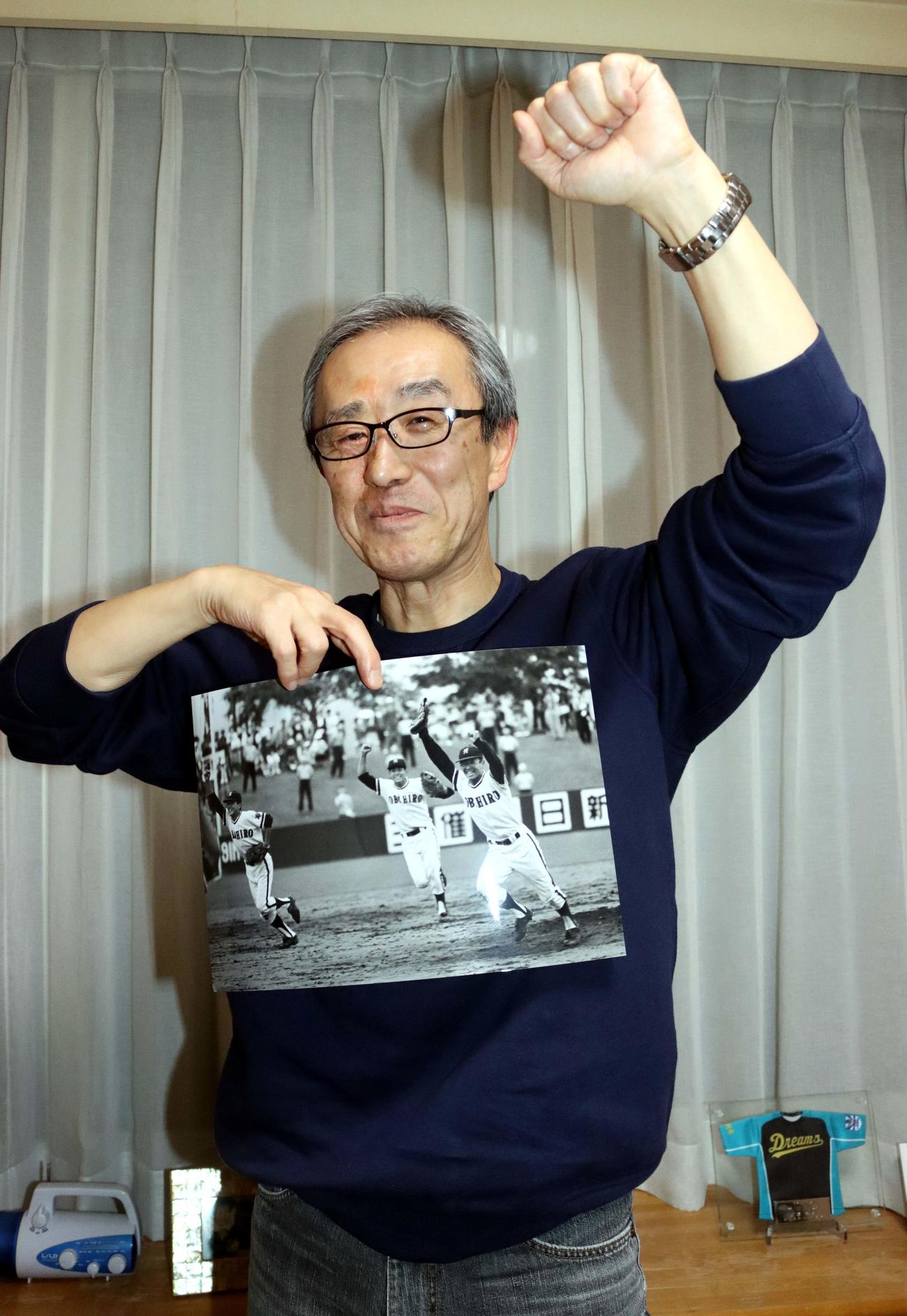82年夏の北北海道大会優勝時の写真を手に、左腕を掲げる帯広農元エースの加藤さん。写真右端が当時の同氏