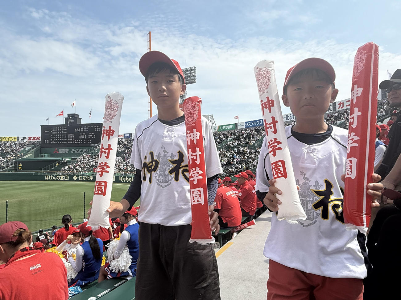 神村学園（鹿児島）の学園カラー・赤をあしらったチアスティックバルーンと、キャップを被って応援する、同校の小田大介監督長男の喜大（はると）さん（12）と次男晃大（あきと）さん（9、写真右）
