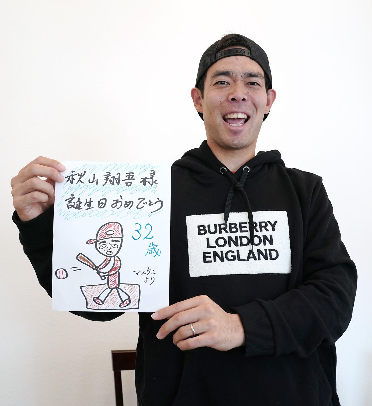 ツインズ前田から誕生日プレゼントとして贈られたパーカを着たレッズ秋山は、同封されていた「マエケン画伯」の似顔絵を笑顔で手にする（本人提供）