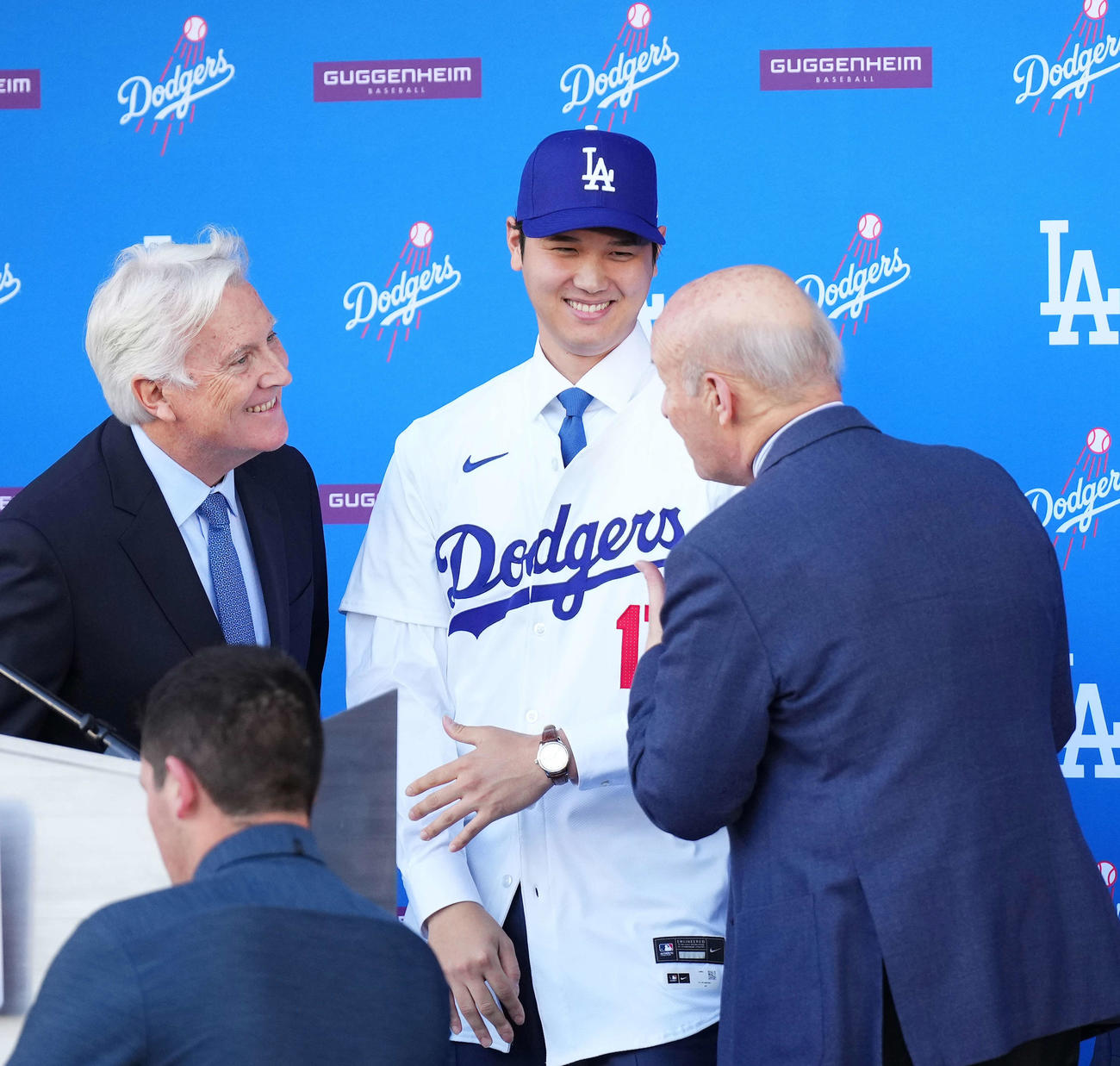 ドジャース入団会見でユニホームに袖を通し笑顔を見せる大谷翔平。左はマーク・ウォルターオーナー、手前右はスタン・カステン球団社長（撮影・菅敏）