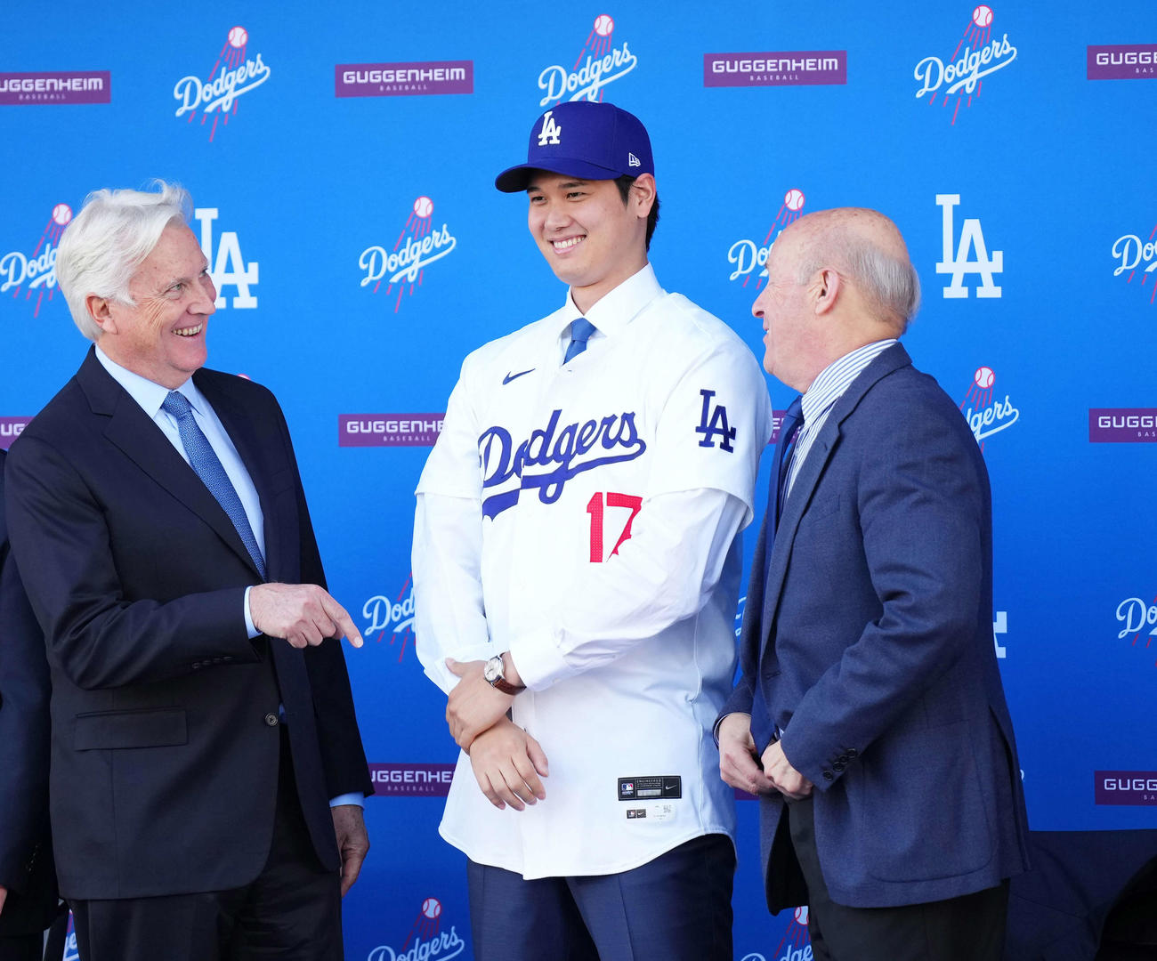 ドジャース入団会見でユニホームに袖を通し笑顔を見せる大谷翔平。左はマーク・ウォルターオーナー、手前右はスタン・カステン球団社長（2023年12月14日撮影）