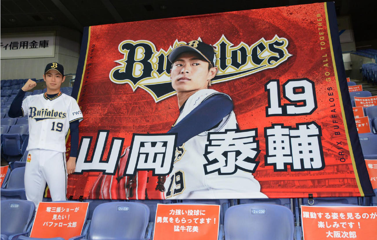 「応援し幕れ！バファローズ自宅応援団」で掲げられる選手横断幕とメッセージペーパーの前でポーズをとるオリックス山岡泰輔（球団提供）