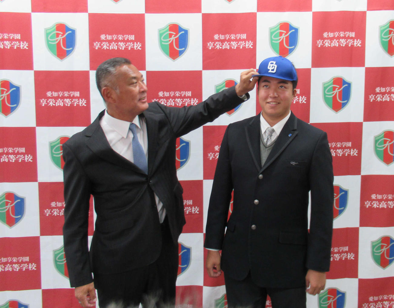 享栄の先輩でもある近藤スカウト（左）から帽子をかぶらせてもらう中日育成2位・上田洸太朗投手