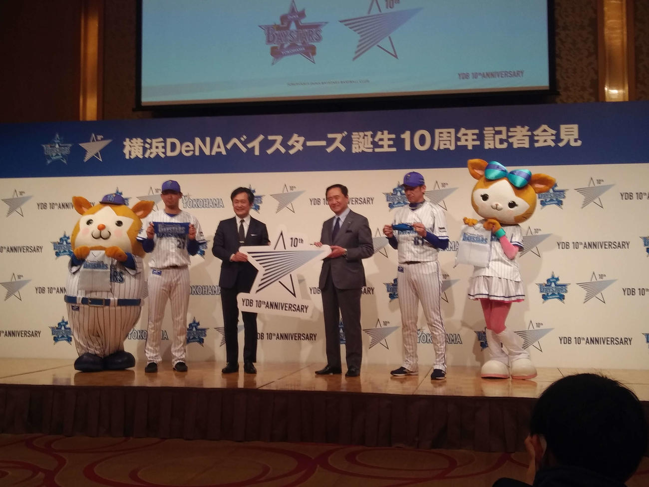 DeNAの球団誕生10周年記者会見。左からプレゼントするナップザックを持つスターマン、プレゼントするタオルを持つ今永昇太、岡村社長、黒岩神奈川県知事、マスクを持つ三浦監督、キララ