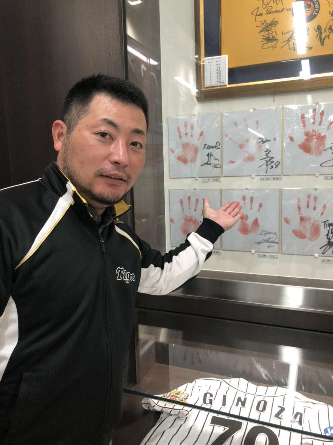 道の駅「ぎのざ」のミニミュージアムに展示中の矢野監督の現役時代の手形を指す、宜野座村観光協会の仲間赴人事務局長（撮影・磯綾乃）