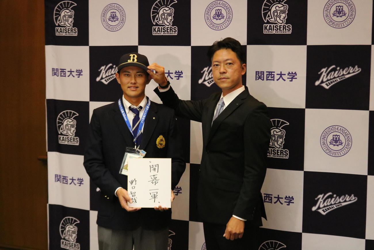 関大の野口智哉内野手は「開幕1軍」と目標を書いた色紙を持ち、オリックス谷口スカウト（右）からオリックスの帽子をかぶせてもらう（撮影・林亮佑）