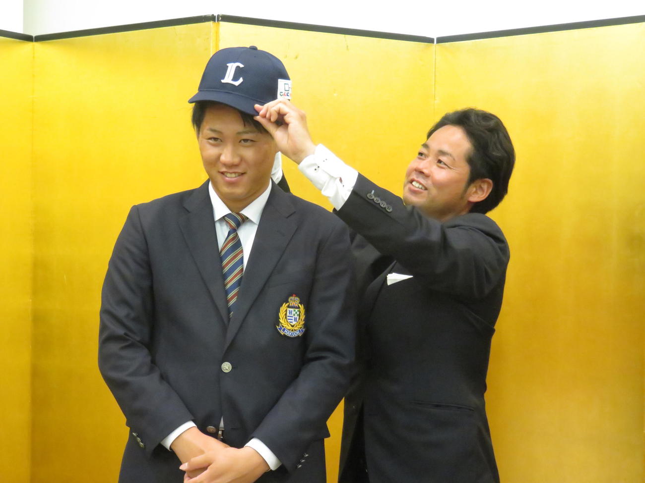 大島裕行スカウト（右）に帽子をかぶらせてもらう身長191センチのドラフト6位・中山誠吾内野手