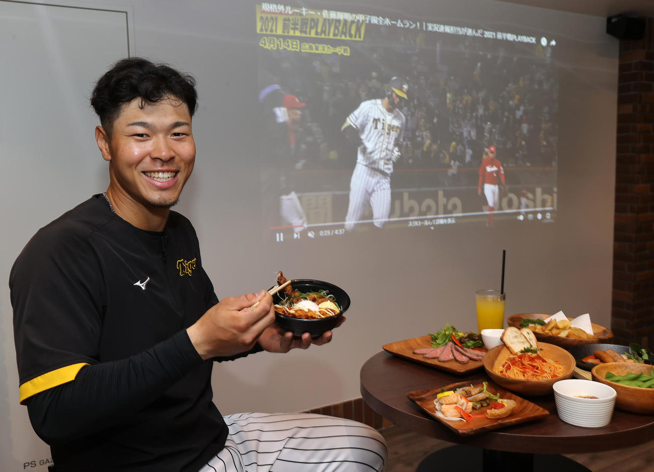 大型プロジェクターの映像をバックに「サトテルのWチキン輝（テリ）ヤキ丼」を試食する阪神佐藤輝（撮影・上山淳一）
