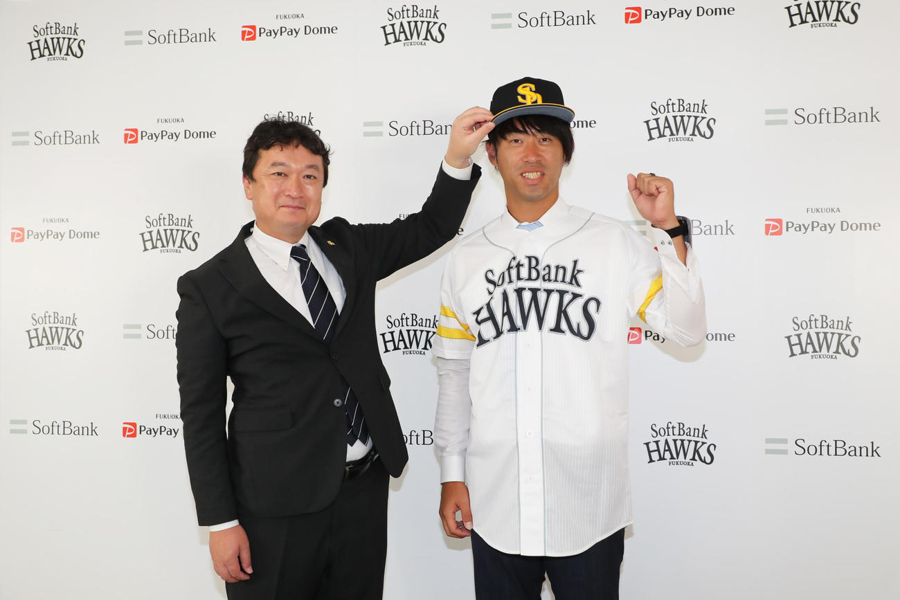入団会見でガッツポーズをする秋吉亮投手。左は三笠取締役GM（外部提供）