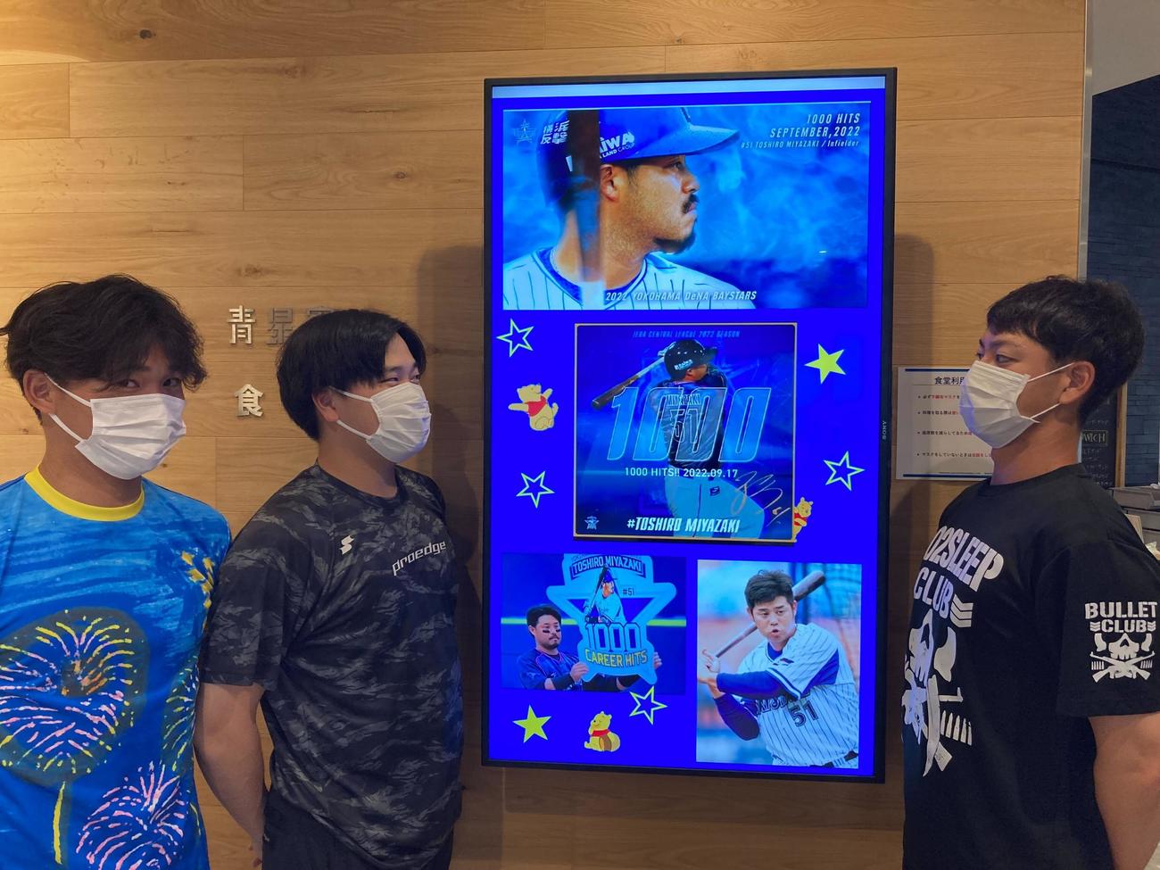 DeNAの球団施設「DOCK」のモニターにある1000安打を達成した宮崎のお祝いページを見つめる、左から高田、加藤、平良（球団提供）