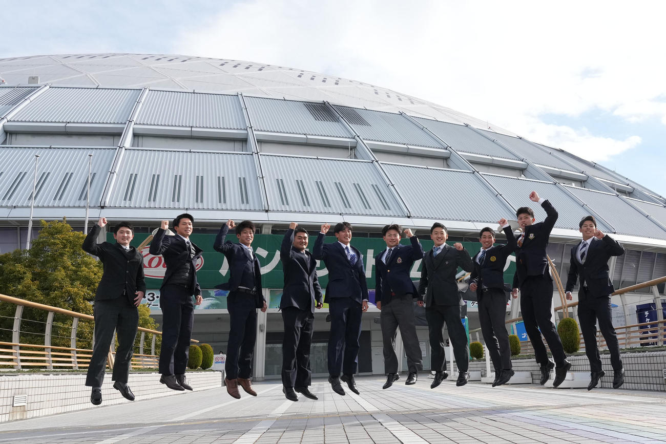 施設を見学し球場を背にジャンプする新入団選手たち、左から野中、福永、浜、森山、仲地、村松、山浅、田中、松山、樋口（撮影・森本幸一）