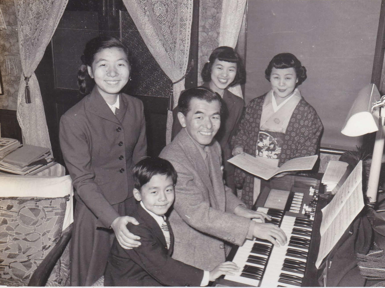 1953年の古関家。自宅でハモンドオルガンの前に座る中央の古関裕而氏、前列の長男正裕さん。後列左から次女紀子さん、長女雅子さん、金子夫人（古関正裕さん提供）