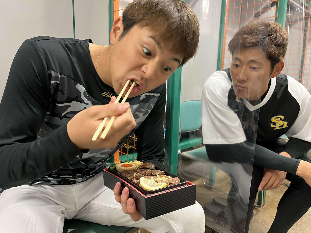 牛乃屋の弁当をほおばるソフトバンク川瀬（左）と羨ましそうに見つめる上林（球団提供）