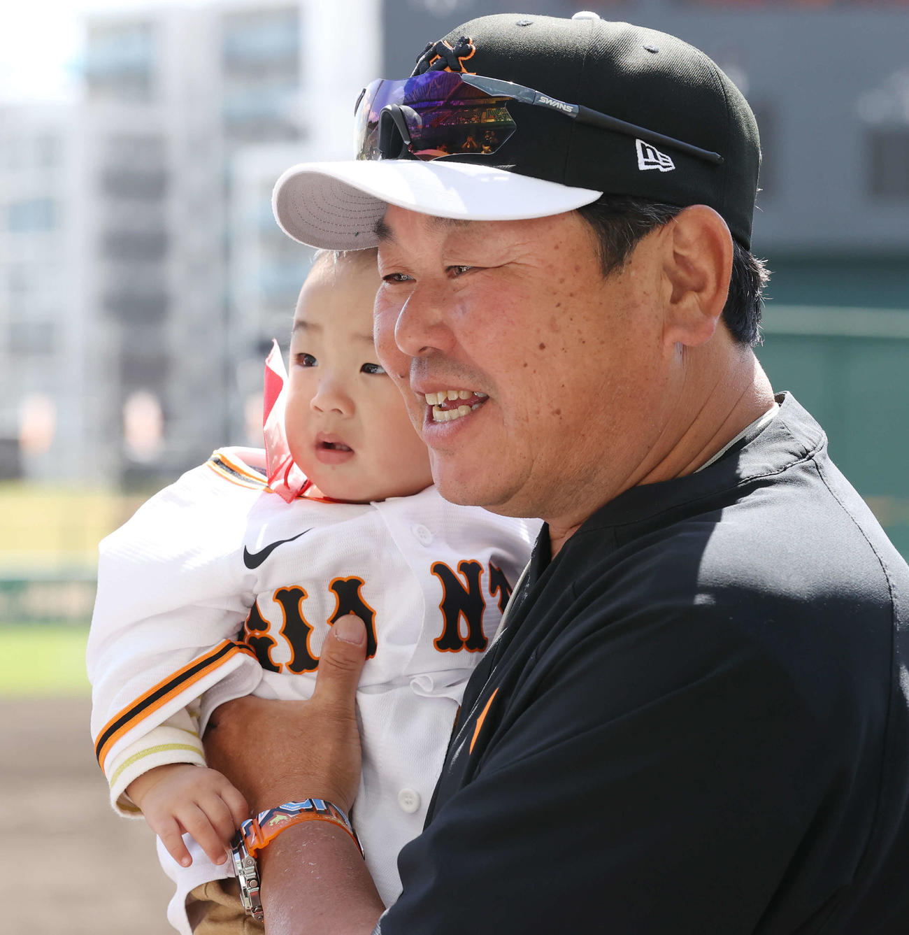 巨人のユニホームを着た赤ちゃんを抱っこし、笑顔の大久保打撃チーフコーチ（撮影・宮地輝）