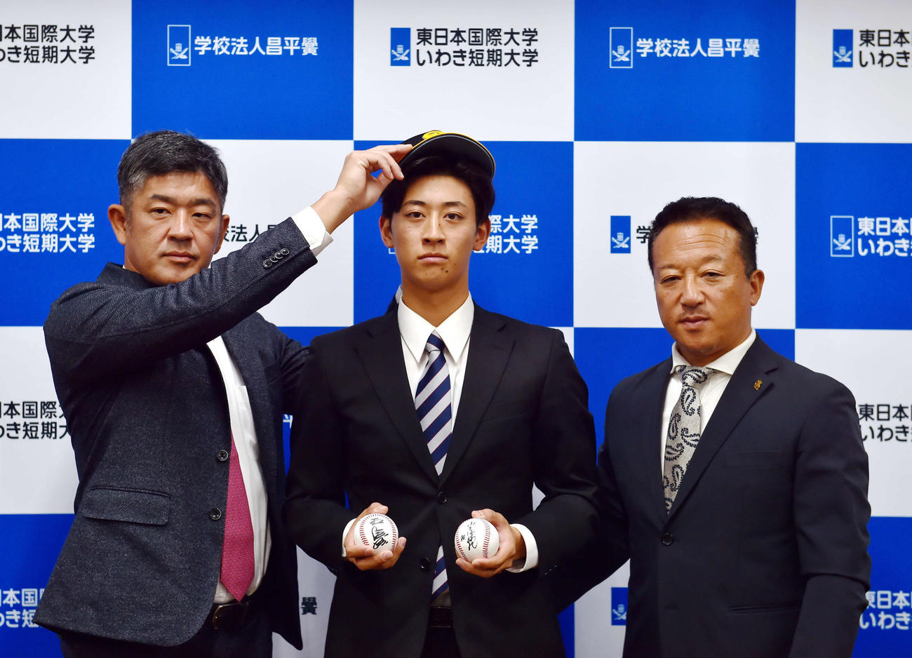 指名あいさつを終え、ソフトバンク福山アマスカウトチーフ（左）に帽子をかぶらせてもらう東日本国際大・大山。右は作山アマスカウトチーフ補佐（撮影・相沢孔志）