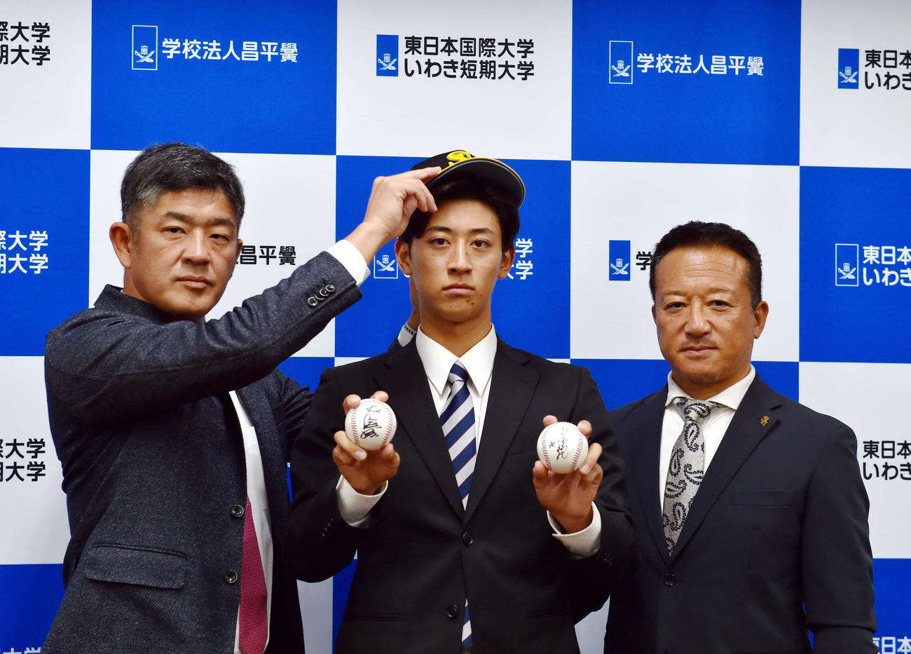 指名あいさつを終え、ソフトバンク福山アマスカウトチーフ（左）に帽子をかぶらせてもらう東日本国際大・大山。右は作山アマスカウトチーフ補佐（撮影・相沢孔志）