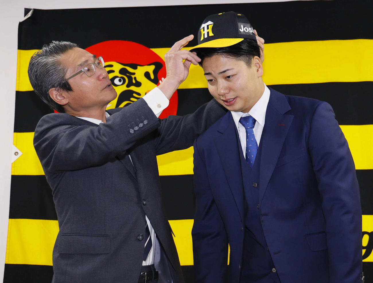 現役ドラフトで阪神に入団し、嶌村球団本部長（左）に帽子をかぶせてもらう漆原（撮影・藤尾明華）