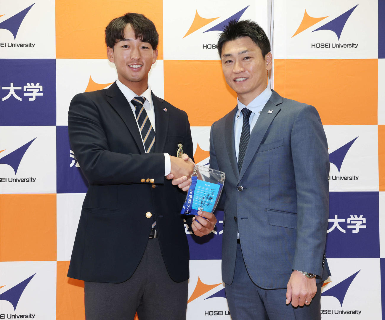 指名あいさつを受け、ヤクルト池山監督の直筆メッセージ入りパスを手に青木GM特別補佐（右）と笑顔で握手するドラフト1位の法大・松下（撮影・宮地輝）