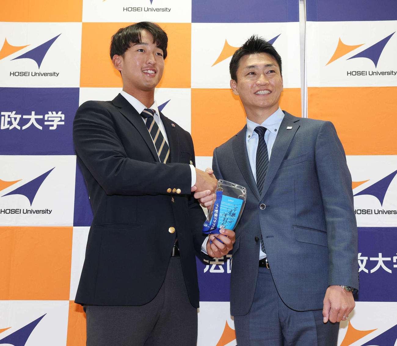 指名あいさつを受け、ヤクルト池山監督の直筆メッセージ入りパスを手に青木GM特別補佐（右）と笑顔で握手するドラフト1位の法大・松下（撮影・宮地輝）