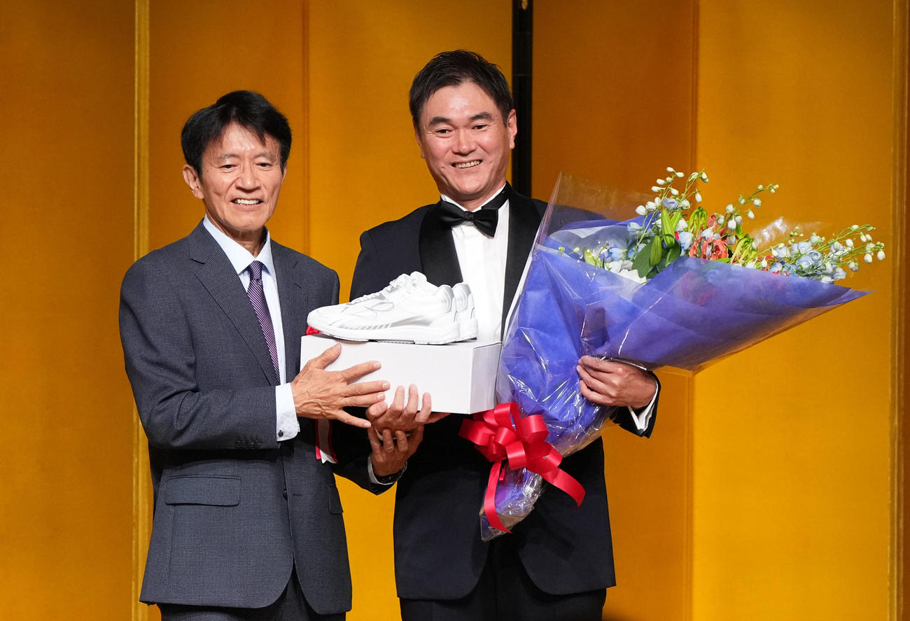 ワールドウイングエンタープライズ代表の小山裕史氏（左）から記念品と花束を受け取り記念撮影に納まる岩瀬仁紀氏（撮影・森本幸一）