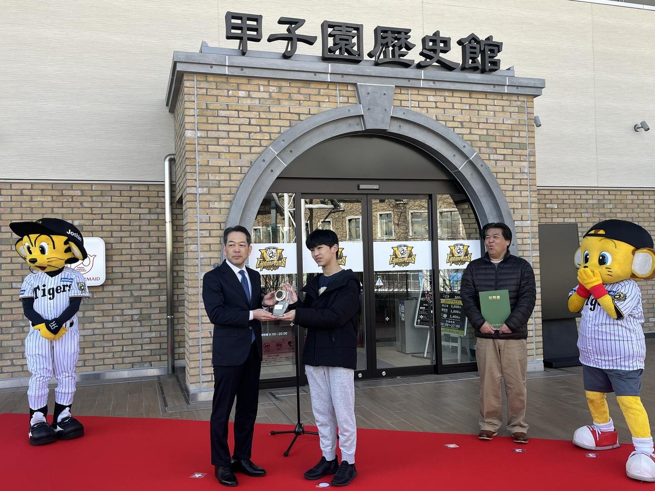「甲子園歴史館　入館者200万人達成セレモニー」に参加し、甲子園歴史館の上戸健司館長から記念品を贈られた田浦大翔さん（中央）。右奥は父の淳一さん（撮影・磯綾乃）