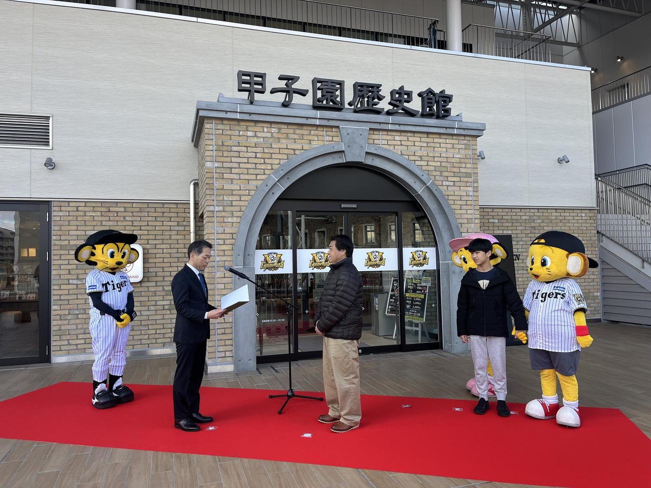 「甲子園歴史館　入館者200万人達成セレモニー」に参加し、甲子園歴史館の上戸健司館長から記念品を贈られた田浦淳一さん（中央）右奥は息子の大翔さん（撮影・磯綾乃）
