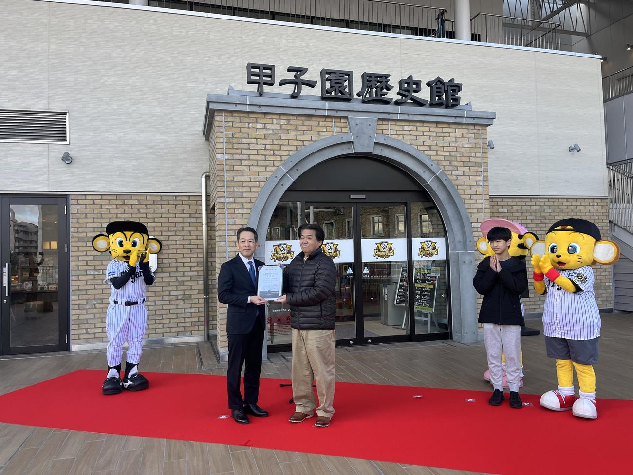「甲子園歴史館　入館者200万人達成セレモニー」に参加し、甲子園歴史館の上戸健司館長から記念品を贈られた田浦淳一さん（中央）右奥は息子の大翔さん（撮影・磯綾乃）