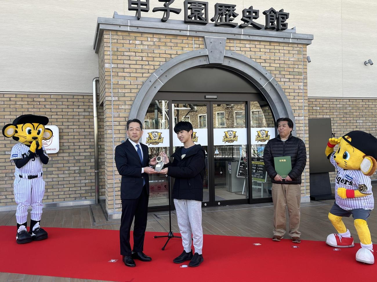 「甲子園歴史館　入館者200万人達成セレモニー」に参加し、甲子園歴史館の上戸健司館長から記念品を贈られた田浦大翔さん（中央）。右奥は父の淳一さん（撮影・磯綾乃）