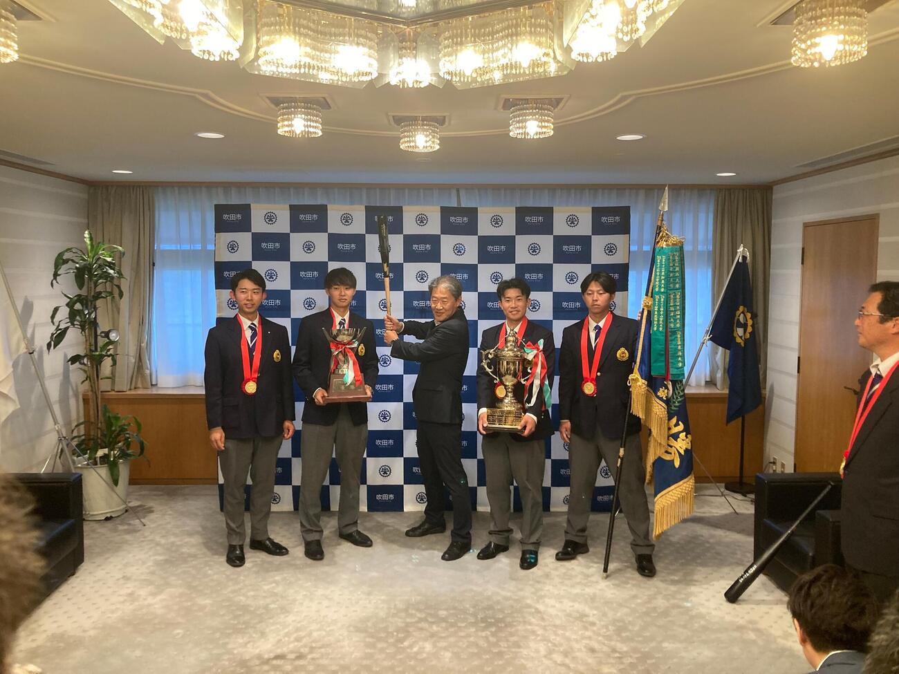 吹田市役所に日本一の報告に訪れた関大野球部メンバー。右から中原海晴副主将、森内大奈主将、バットを構える後藤圭二市長、米沢友翔副主将、大田侑季主務