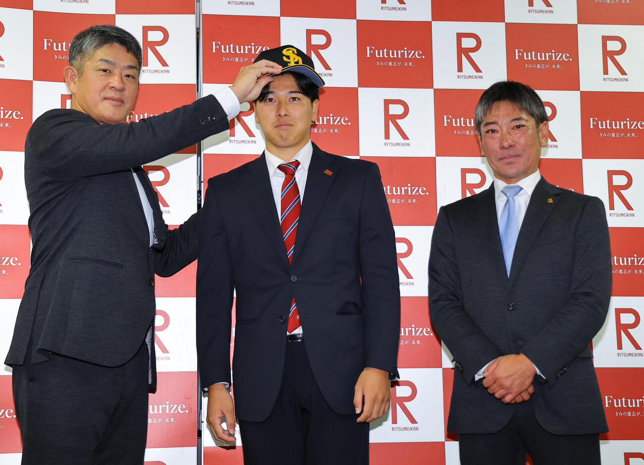 指名あいさつを受けたソフトバンクドラフト2位の立命大・有馬伽久（中央）は福山龍太郎編成育成本部長補佐兼スカウト部アマスカウトチーフ（左）から帽子を被せてもらう。右は稲嶺誉スカウト（撮影・上田博志）