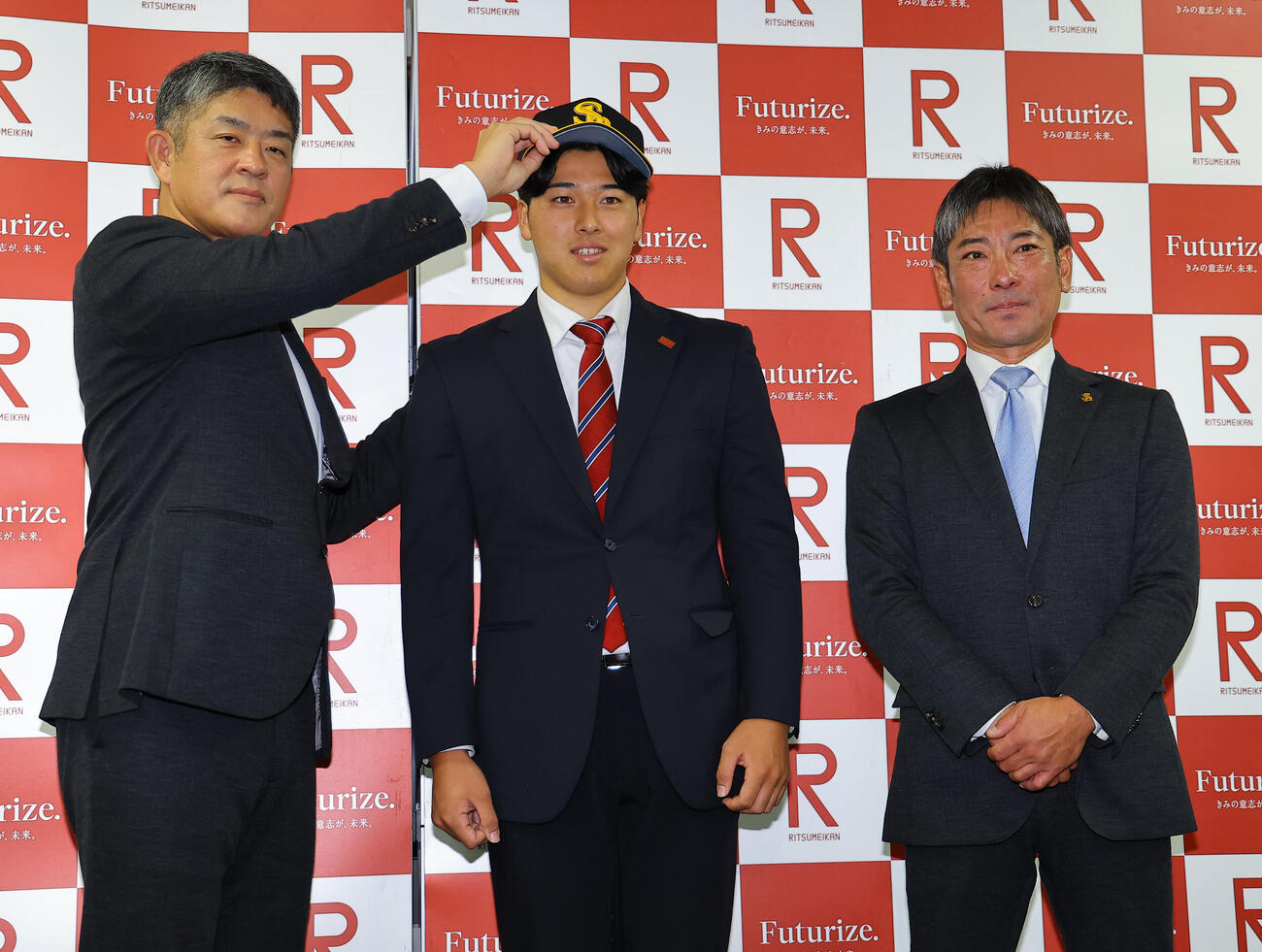 指名あいさつを受けたソフトバンクドラフト2位の立命大・有馬伽久（中央）は福山龍太郎編成育成本部長補佐兼スカウト部アマスカウトチーフ（左）から帽子を被せてもらう。右は稲嶺誉スカウト（撮影・上田博志）