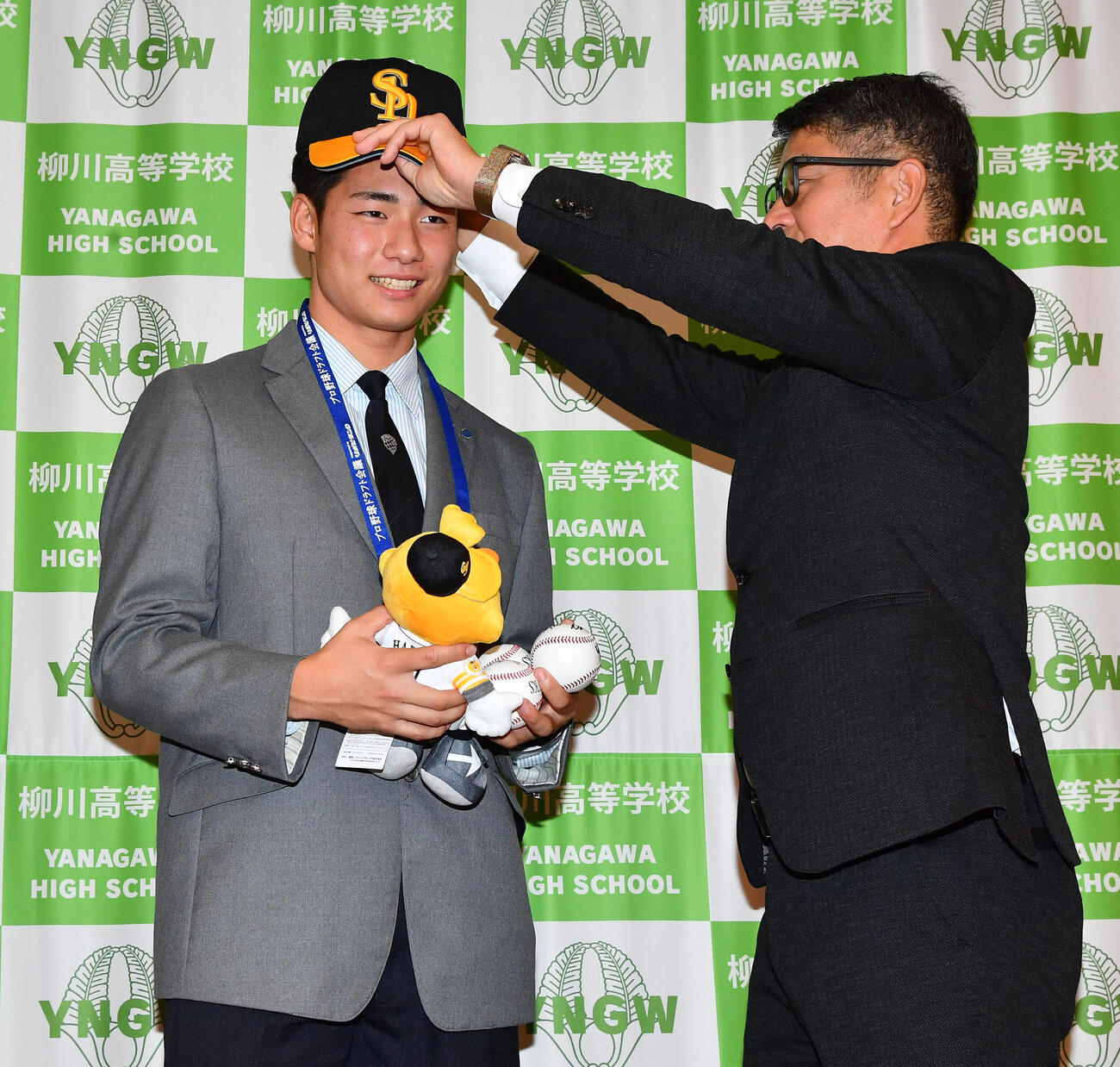 ソフトバンクからドラフト6位で指名され、福山龍太郎編成育成本部長補佐兼アマスカウトチーフ（右）から帽子をかぶせてもらう小坪碧人（撮影・岩下翔太）