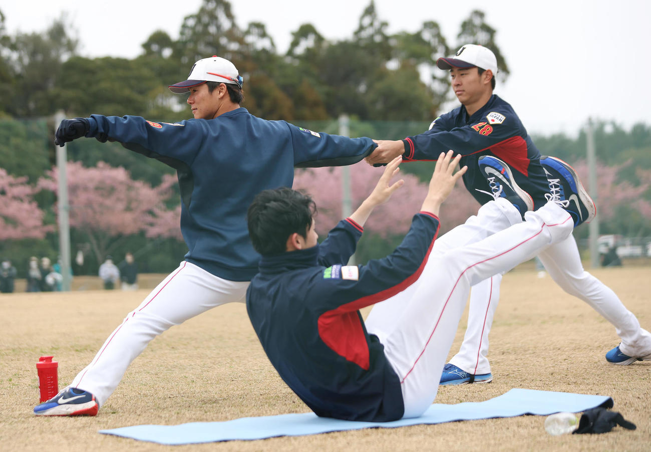 投球の姿勢で高橋宏（右）に腕を引いてもらう山本。手前はトレーニングする佐々木（撮影・前田充）