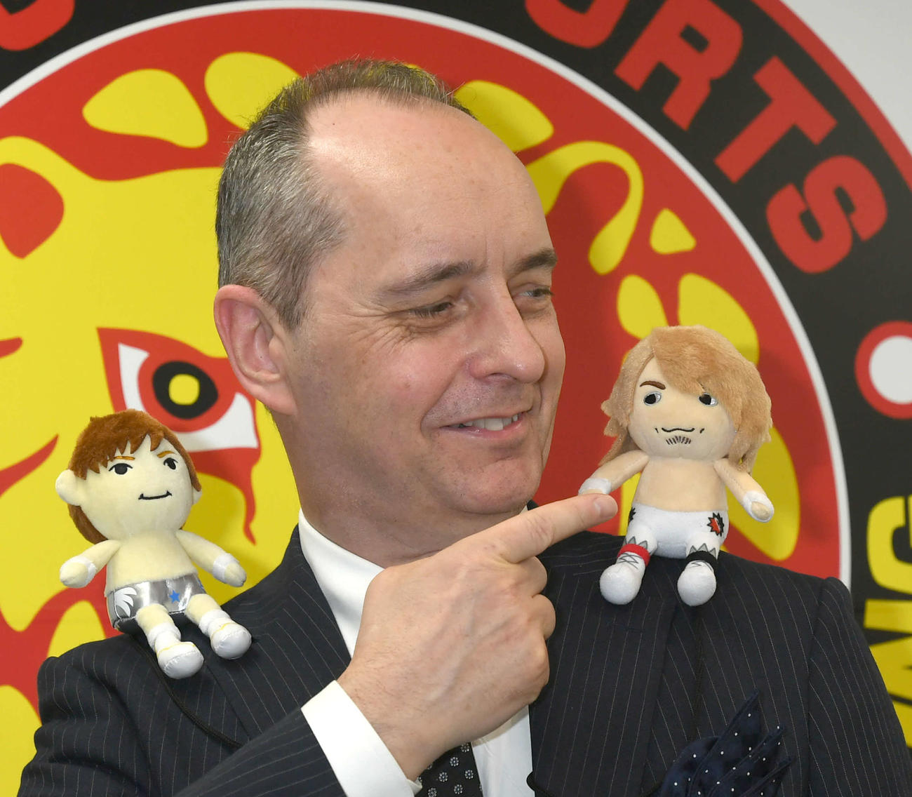 選手のマスコット人形を肩に付けて、笑顔でポーズをとる新日本プロレスのメイ社長