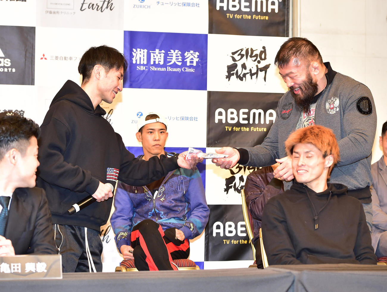 前日会見、ほかの対戦カードの選手が挑発で出してきたサラダチキンを「なら自分が」と名乗り出て受け取るサトシ・イシイ（右）（撮影・清水貴仁）