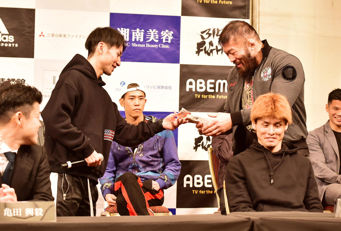 前日会見、ほかの対戦カードの選手が挑発で出してきたサラダチキンを「なら自分が」と名乗り出て受け取るサトシ・イシイ（右）（撮影・清水貴仁）
