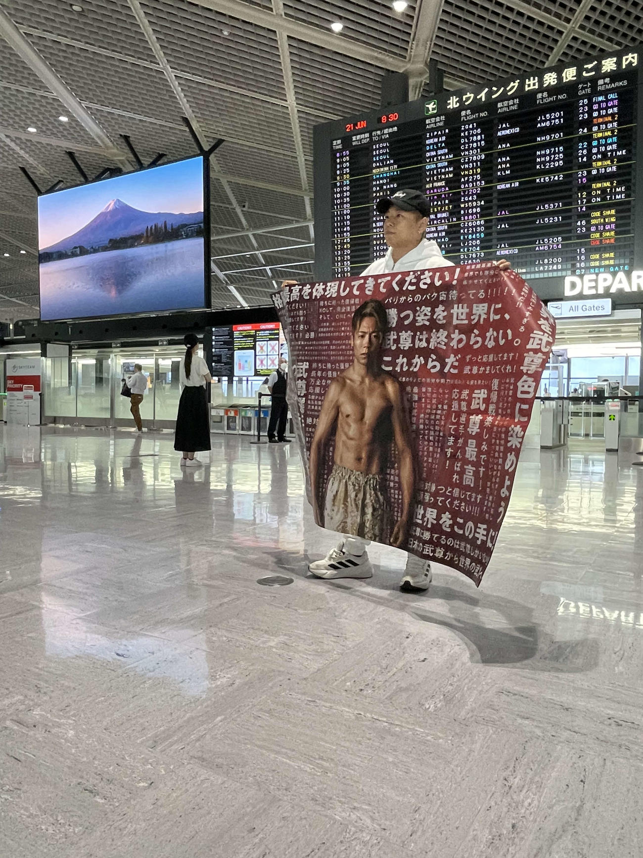奥のビジョンで富士山が映し出される中、復帰戦の地フランスへ渡航前に、出発ゲートでファンからの横断幕を手に気合の入った表情を見せる武尊