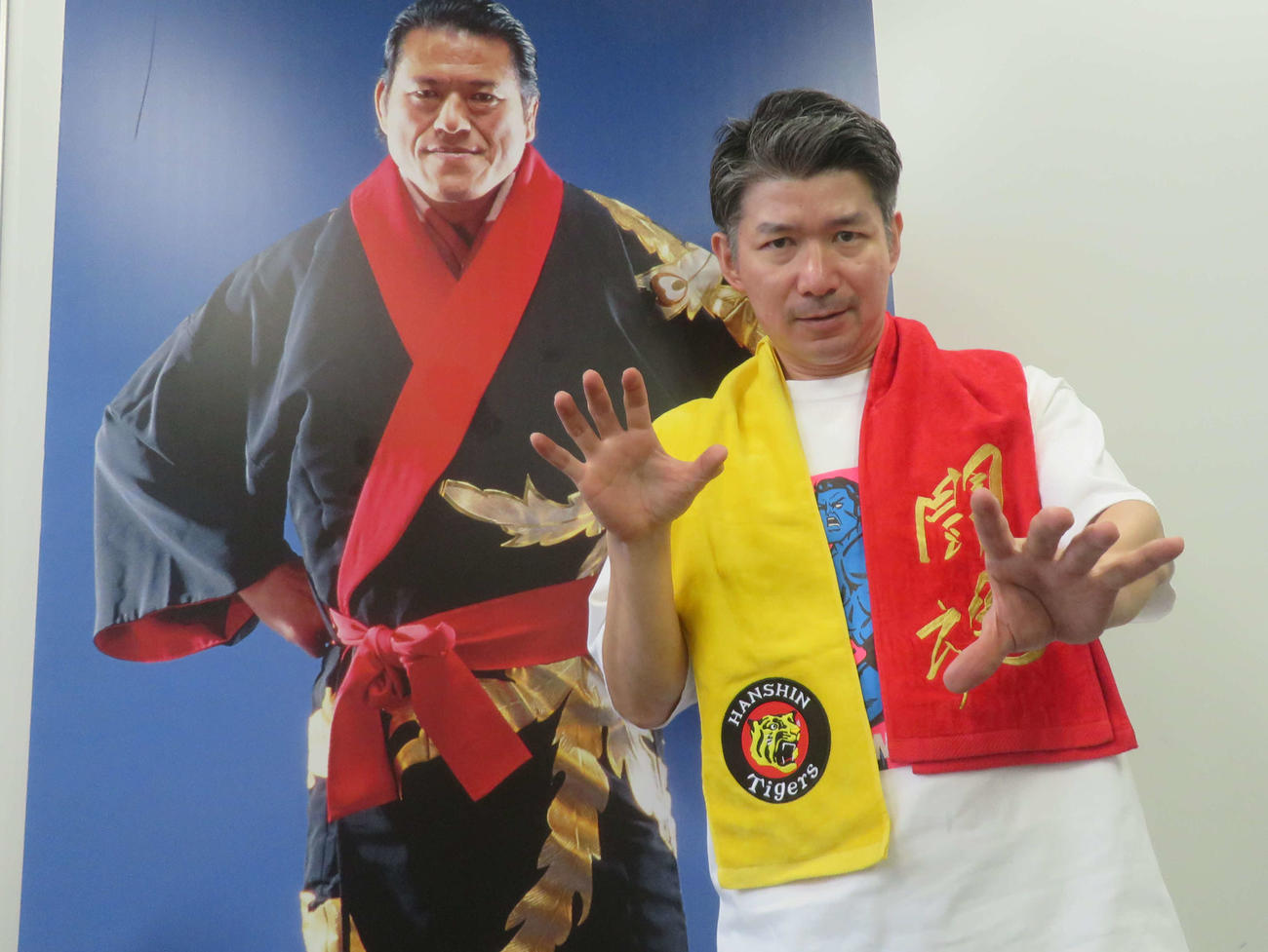 猪木展で販売される阪神タイガースとアントニオ猪木のコラボタオルマフラーを巻き、猪木展のみどころを語るIGFの宇田川強取締役