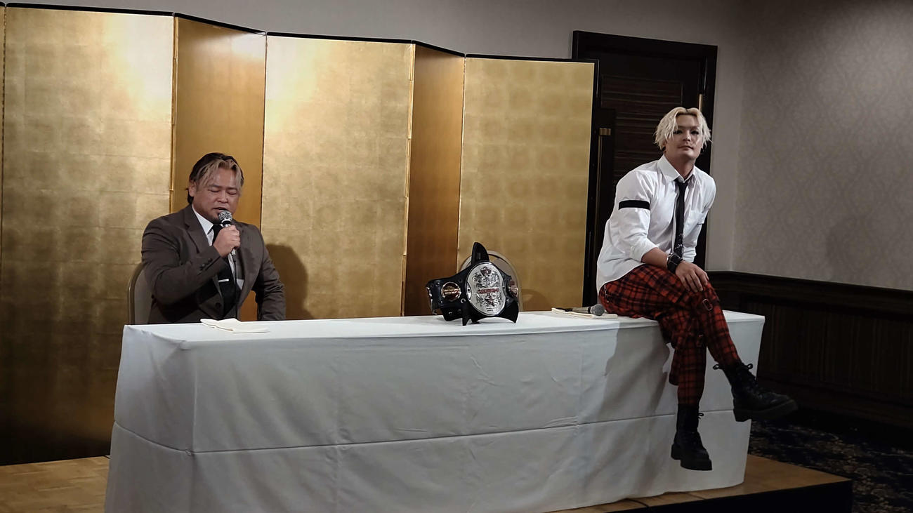 カズ・ハヤシが話す隣で足を組んで机の上に座るいつもと変わらぬライジングHAYATO（右）