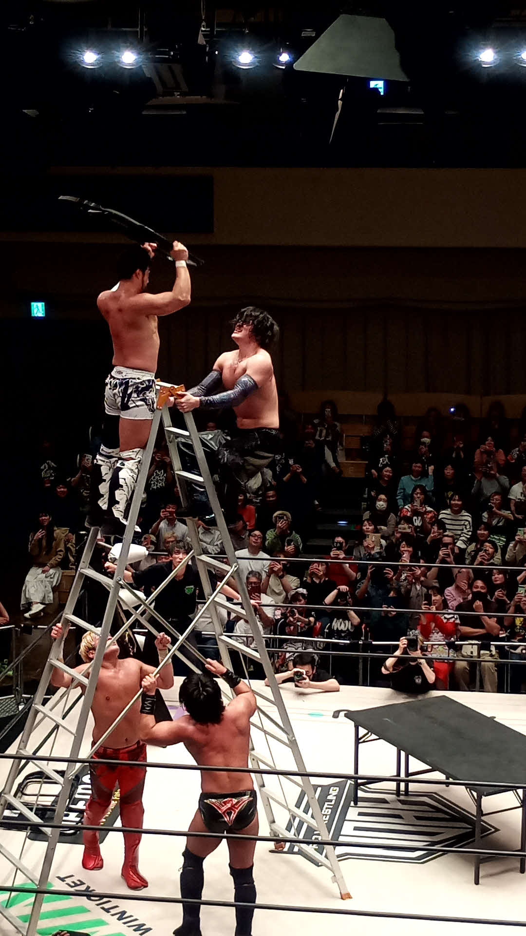 巨大脚立の上からいすでOZAWAをリング上の長机へたたき落としたKENTA