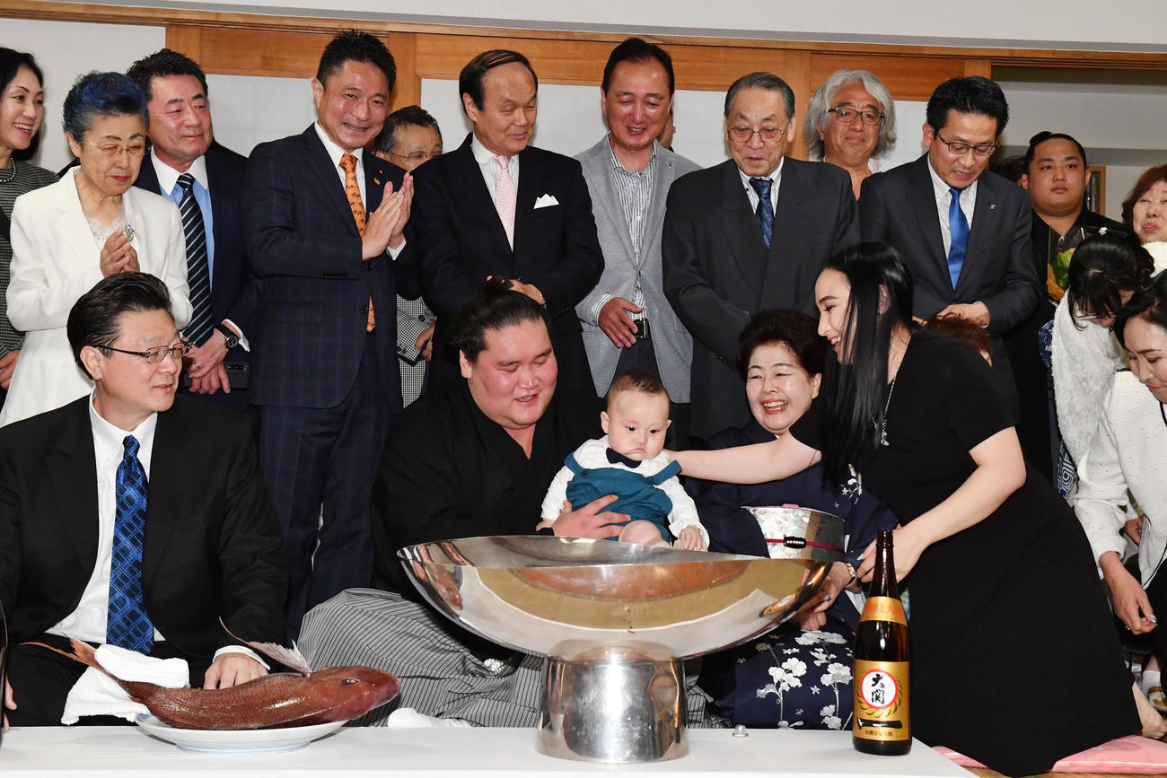 夏場所千秋楽、伊勢ケ浜部屋での祝賀会でツェグメド・ドルジハンド夫人（右）から手渡された長男・テムジン君を抱く照ノ富士。前列左は師匠の伊勢ケ浜親方、前列右から2人目はおかみさんの淳子夫人（2023年5月28日撮影）
