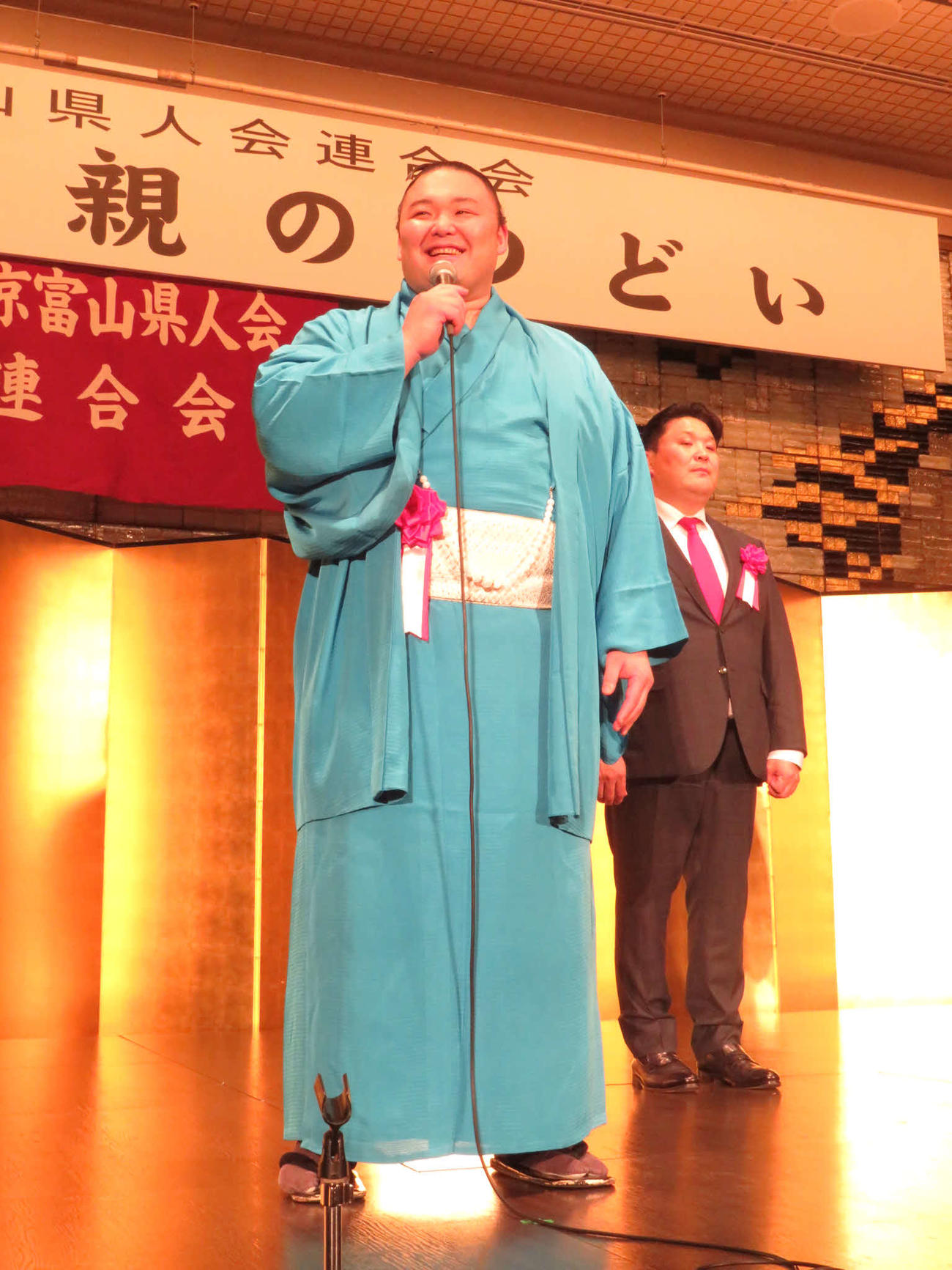 東京富山県人会連合会の「懇親のつどい」に出席し、壇上で笑顔であいさつした朝乃山（左）。右は高砂親方