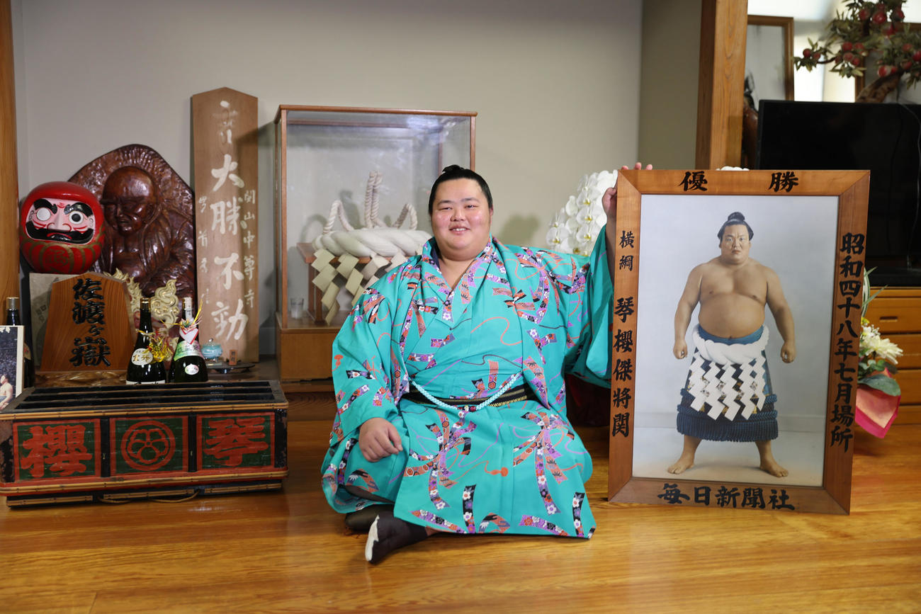 大相撲初場所千秋楽から一夜明け、祖父で先代佐渡ケ嶽親方の元横綱琴桜が使用していた明け荷と優勝額の間に座り笑顔の琴ノ若（代表撮影）