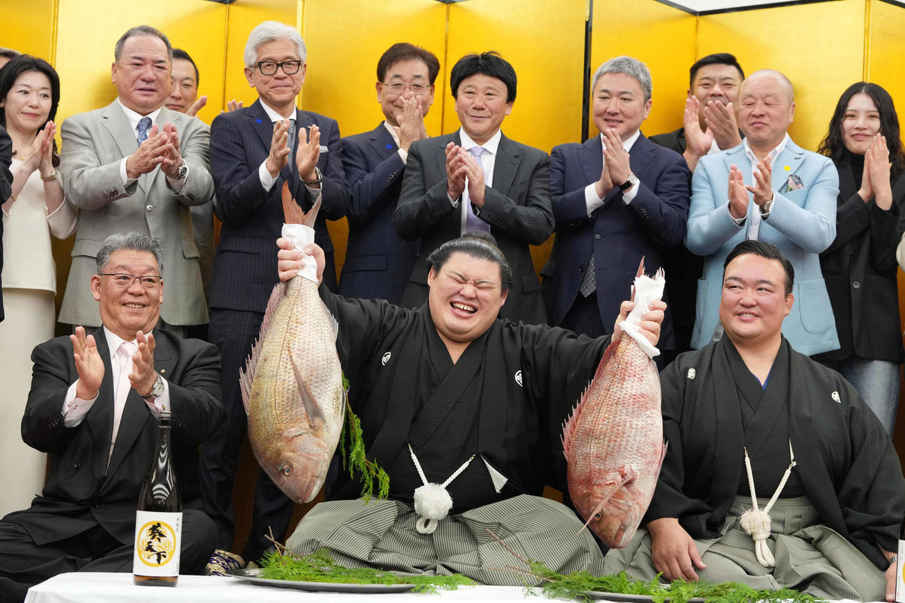 関係者の祝福を受けながら両手でお祝いのタイを持ち上げる大の里。前列左は二所ノ関部屋大東後援会の橋本正幸会長、右は二所ノ関親方（撮影・小沢裕）