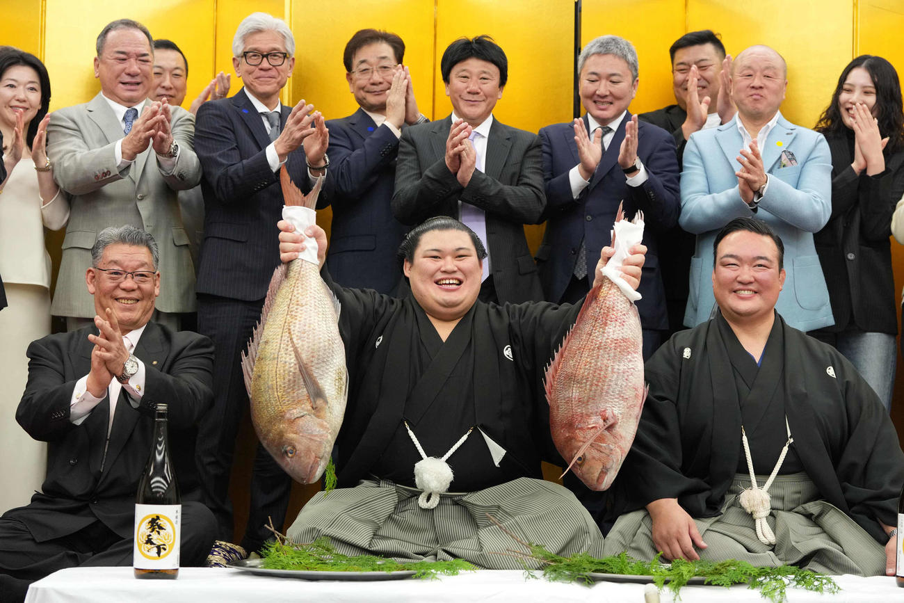 関係者の祝福を受けながら両手でお祝いのタイを持ち上げる大の里。前列左は二所ノ関部屋大東後援会の橋本正幸会長、右は二所ノ関親方（撮影・小沢裕）