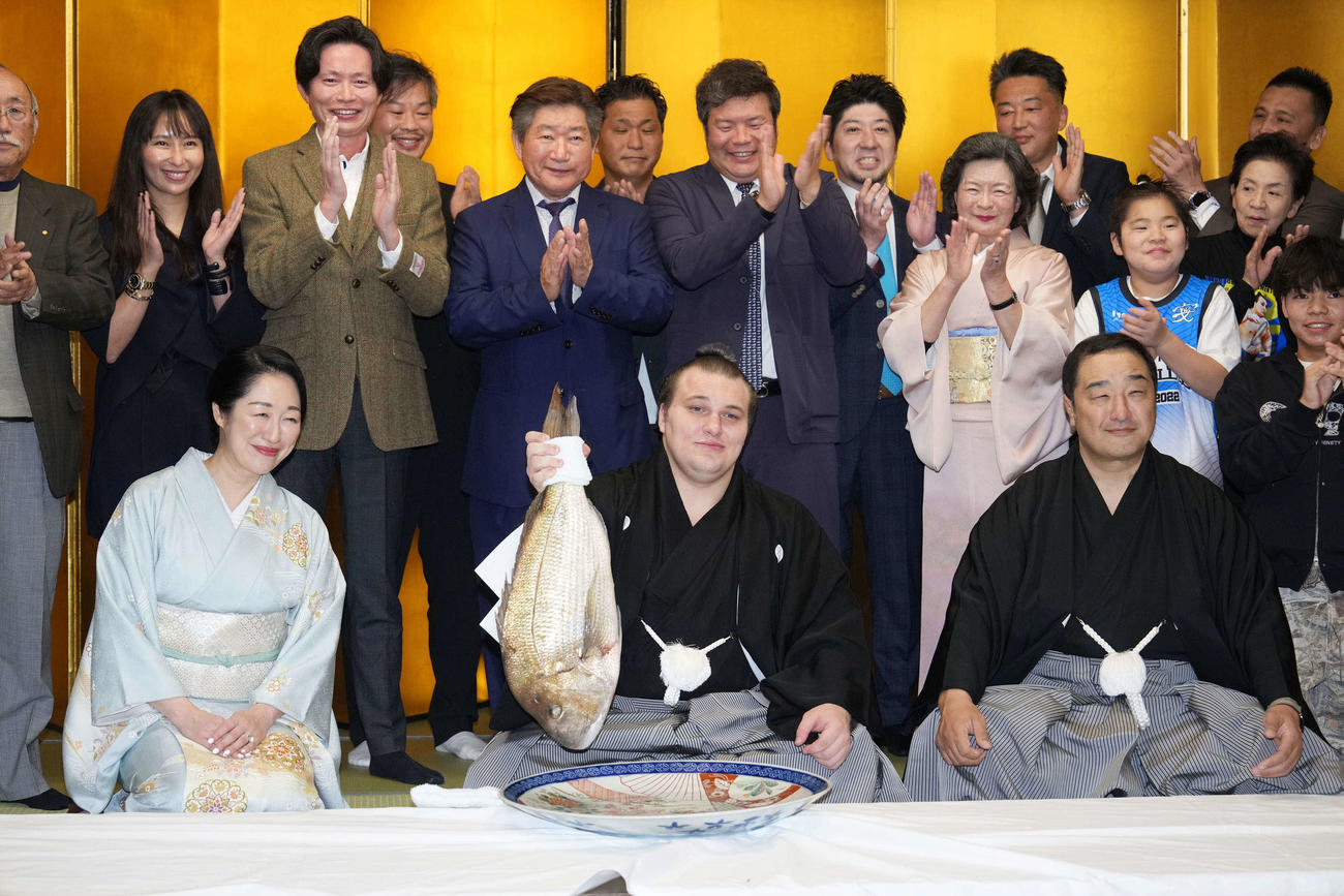 千秋楽パーティーで師匠の安治川親方（右）、おかみの絵莉夫人、関係者の祝福を受けながらお祝いのタイを持ち上げる安青錦（撮影・小沢裕）