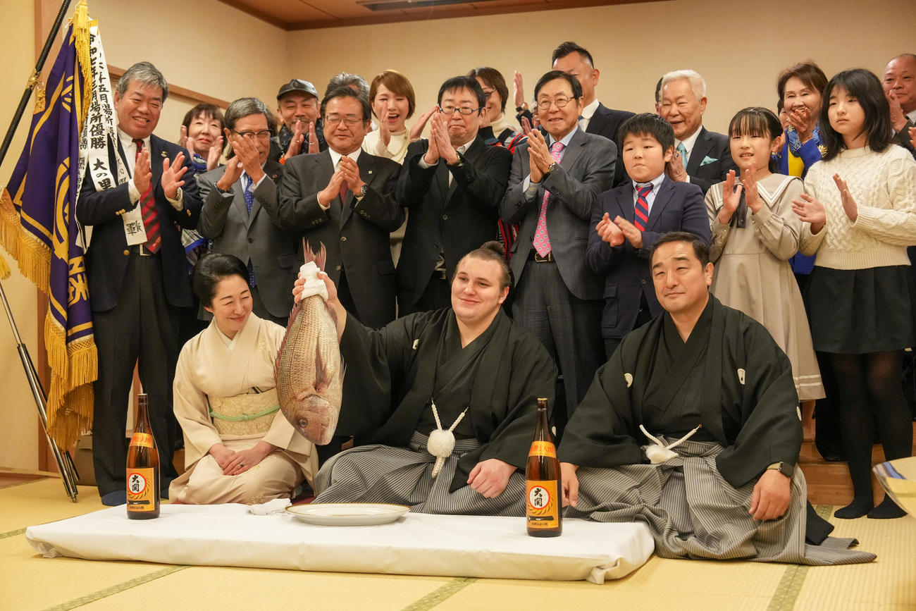 関係者の祝福を受けながらお祝いの鯛を持ち上げる安青錦。右は師匠の安治川親方、左はおかみの絵莉さん（撮影・小沢裕）